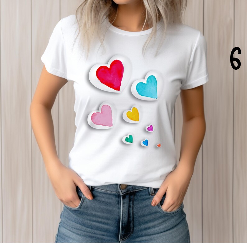 May include: White t-shirt featuring a watercolor design of colorful hearts in red, blue, pink, yellow, and green. The hearts have a white outline and a 3D effect. The shirt is worn by a person in blue jeans.