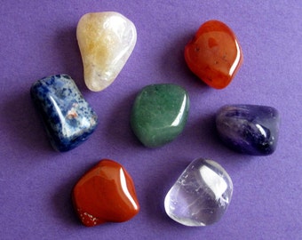 May include: Seven smooth, polished stones in various colours, including clear, yellow, green, blue, red, orange, and purple, arranged on a purple background. These stones are often used in crystal healing and meditation.