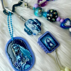 Puede incluir: Una colección de llaveros y accesorios con cuentas en tonos azules, morados y plateados. Los llaveros presentan imágenes de una novia. Los accesorios con cuentas incluyen cuentas de varios tamaños y formas.