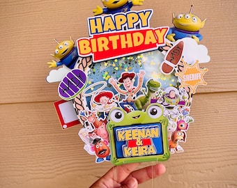 Könnte beinhalten: Ein mehrschichtiger Tortenaufleger im Toy Story-Design mit drei grünen Aliens oben, einem zentralen Schüttel-Element mit goldenem Glitzer und verschiedenen Charakter-Ausschnitten wie Woody, Buzz Lightyear und Rex. Der Text lautet HAPPY BIRTHDAY und KEENAN & KEIRA auf einem grünen, froschförmigen Rahmen.