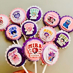 May include: A collection of decorative cupcake toppers with various designs. Each topper features a circular image with a scalloped edge, mounted on a white stick. The designs include cartoon characters, names, and the text "HUNTRAX".