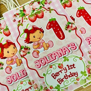 May include: Pink and white checkered party bags with a strawberry theme. Each bag features a cartoon girl, the name "Joliana's", and the text "Berry 1st Birthday". The bags have white string handles.