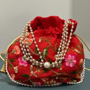 Może przedstawiać: Czerwona torebka ze sznurkiem z haftem kwiatowym w kolorze różowym, pomarańczowym i zielonym. Torebka jest ozdobiona złotymi cekinami i warstwowym naszyjnikiem z pereł. Sznurek jest złoty, a torebka obszyta perłami.