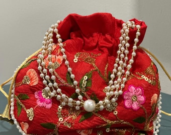 May include: A red drawstring pouch with floral embroidery in pink, orange, and green. The pouch is adorned with gold sequins and a layered pearl necklace. The drawstring is gold, and the pouch is trimmed with pearls.