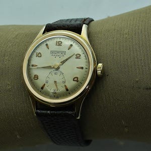Può includere: Un orologio da polso vintage color oro con quadrante color crema e cinturino in pelle nera. Il quadrante dell'orologio presenta numeri neri, un quadrante secondario e la scritta "Pontiac Nageur". L'orologio è esposto su una superficie verde.
