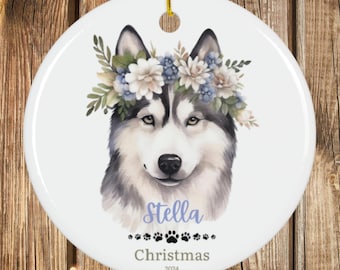 May include: A white ceramic Christmas ornament featuring a watercolor portrait of a Husky dog wearing a floral crown. The ornament is personalized with the name "Stella", paw prints, and the text "Christmas 2024". A gold string is attached for hanging.