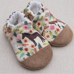 May include: A pair of baby booties with a floral and horse print. The booties are made of a soft, white fabric with a brown suede sole.