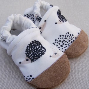 May include: A pair of white baby booties with a hedgehog print and brown suede soles.