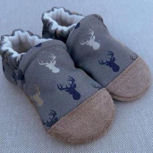 May include: A pair of gray baby booties with a deer head pattern in white and navy blue. The soles are a light brown suede, and the interior is lined with white fabric. These soft-soled shoes are designed for infants.
