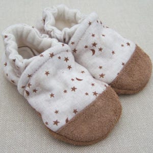 May include: A pair of baby booties with a white fabric upper adorned with brown stars and crescent moons. The toe area is a soft brown suede-like material. The booties have an elasticated opening.