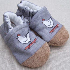 May include: A pair of gray baby booties with a brown suede bottom. The booties feature a white chicken on a skateboard graphic.
