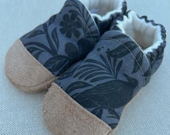 Può includere: Un paio di scarpine per bambini con un motivo floreale e di uccelli blu scuro. Le scarpine hanno la punta in pelle scamosciata marrone chiaro e un bordo bianco a balze. Il design presenta un corvo ed elementi floreali.