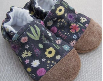 Può includere: Un paio di scarpine per neonati con stampa floreale su sfondo blu scuro. Le scarpine hanno le punte in camoscio marrone e sono foderate con tessuto bianco.