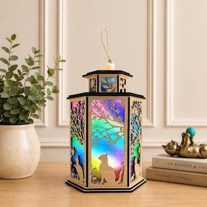 May include: A hexagonal wooden lantern featuring laser-cut silhouettes of cats and trees. The panels are backlit with a vibrant, multi-colored gradient light. The lantern has a black frame and a natural wood finish, topped with a jute rope handle.