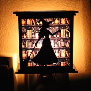 Op de afbeelding: Zwart nachtlampje met een heksensilhouet met een bezem, tegen een achtergrond van een boekenplank vol boeken en potten. Het licht geeft een warme gloed, waardoor een gezellig Halloween- of fantasiethema ontstaat.