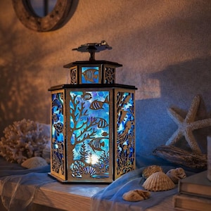 Puede incluir: Una linterna hexagonal de madera con paneles cortados a láser que representan una escena submarina con peces, corales y vida marina. La linterna emite una luz azul brillante desde el interior, resaltando los recortes sobre el marco de madera.