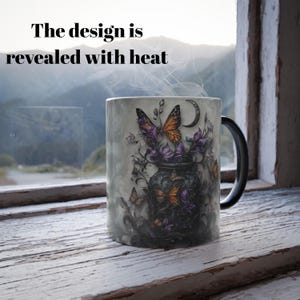 May include: A black and white heat-sensitive mug with a design of a jar filled with purple flowers and orange butterflies. The text "The design is revealed with heat" is visible above the mug.