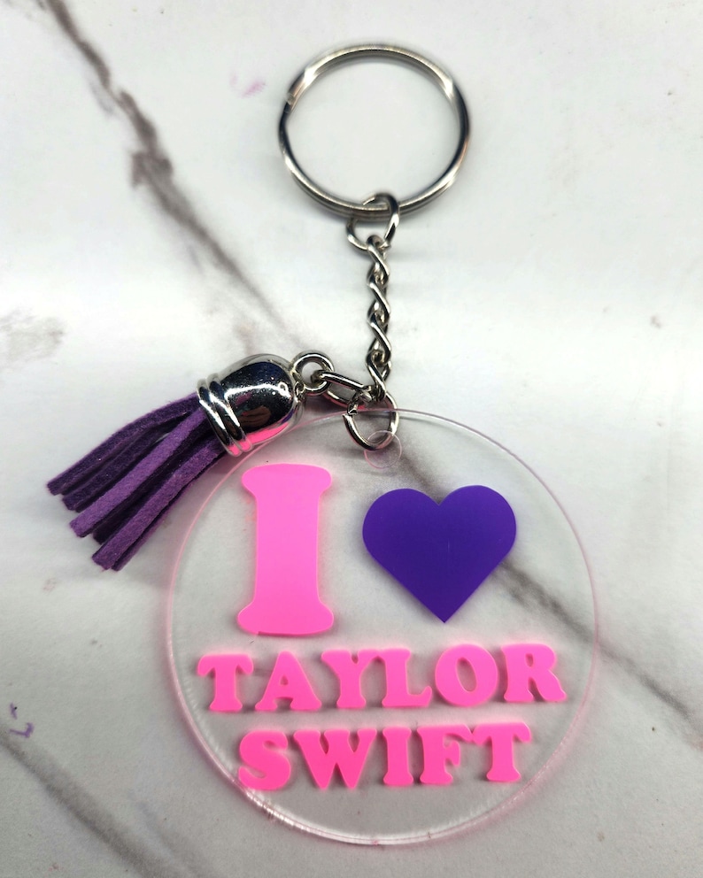 Puede incluir: Un llavero de acr&iacute;lico transparente con un dise&ntilde;o rosa de "I love Taylor Swift". El llavero tiene un coraz&oacute;n morado y un borla morado.