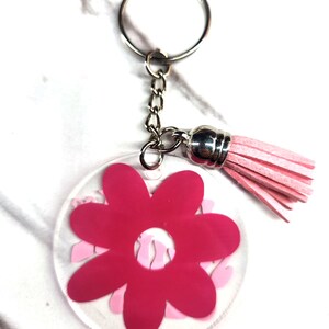 Pode incluir: Um chaveiro com a forma de uma flor rosa com um fundo de acr&iacute;lico transparente e um tassel rosa. O chaveiro est&aacute; ligado a um anel de chave prateado.