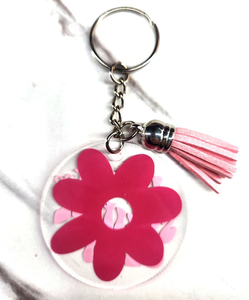 Pode incluir: Um chaveiro com a forma de uma flor rosa com um fundo de acr&iacute;lico transparente e um tassel rosa. O chaveiro est&aacute; ligado a um anel de chave prateado.