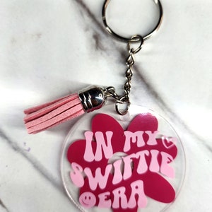 Pode incluir: Chaveiro de acr&iacute;lico rosa e vermelho em forma de flor com o texto "In My Switie Era" com um cora&ccedil;&atilde;o rosa e um rosto sorridente. O chaveiro tem um anel prateado e um tassel rosa.