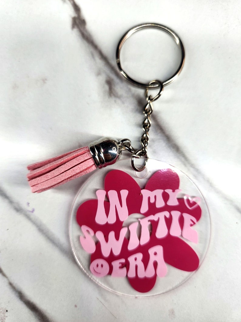 Pode incluir: Chaveiro de acr&iacute;lico rosa e vermelho em forma de flor com o texto "In My Switie Era" com um cora&ccedil;&atilde;o rosa e um rosto sorridente. O chaveiro tem um anel prateado e um tassel rosa.