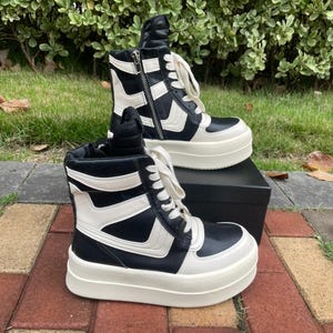 May include: Black and white high-top sneakers with a thick, off-white sole. The sneakers feature a side zipper and white laces. The design includes white angular accents on the black upper. The shoes are displayed on a black box and red brick surface.