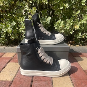 May include: Black high-top sneakers with a white toe cap and sole. The sneakers have a side zipper and thick, light-colored laces. The shoes are displayed on a gray box against a backdrop of green foliage and a brick surface.