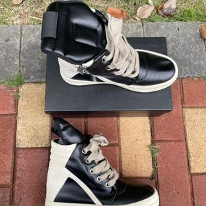 May include: Black and white high-top sneakers with a unique design. The sneakers feature a black leather upper with white accents, a zipper detail, and thick, off-white soles. The laces are a light beige color. The sneakers are displayed on a black box.