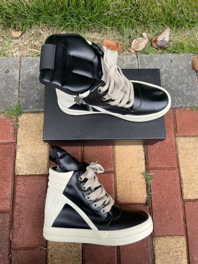 May include: Black and white high-top sneakers with a unique design. The sneakers feature a black leather upper with white accents, a zipper detail, and thick, off-white soles. The laces are a light beige color. The sneakers are displayed on a black box.