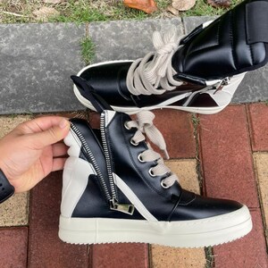 May include: Black and white high-top sneakers with a side zipper and cream-colored laces. The sneakers have a thick, white sole and a contrasting white panel. The shoes are made of leather and are designed for casual wear.