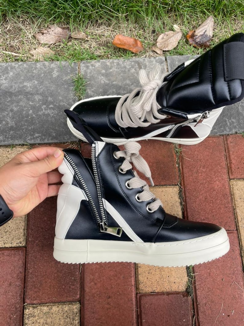 May include: Black and white high-top sneakers with a side zipper and cream-colored laces. The sneakers have a thick, white sole and a contrasting white panel. The shoes are made of leather and are designed for casual wear.