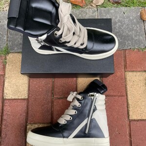 May include: Black and white high-top sneakers with a side zipper and thick, off-white soles. The sneakers have light-colored laces and are displayed on a black box. The design includes a white star-like accent on the side.