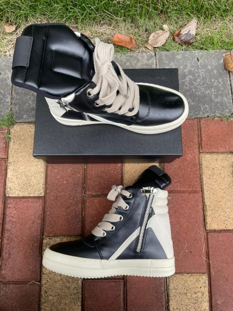 May include: Black and white high-top sneakers with a side zipper and thick, off-white soles. The sneakers have light-colored laces and are displayed on a black box. The design includes a white star-like accent on the side.
