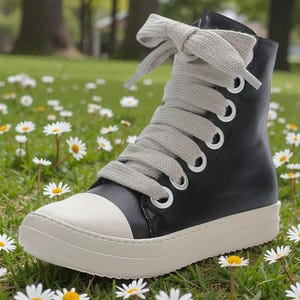 May include: Black high-top sneaker with a white toe cap and thick, off-white sole. The shoe features large, gray laces and silver eyelets. The sneaker is set against a backdrop of green grass and small white daisies.