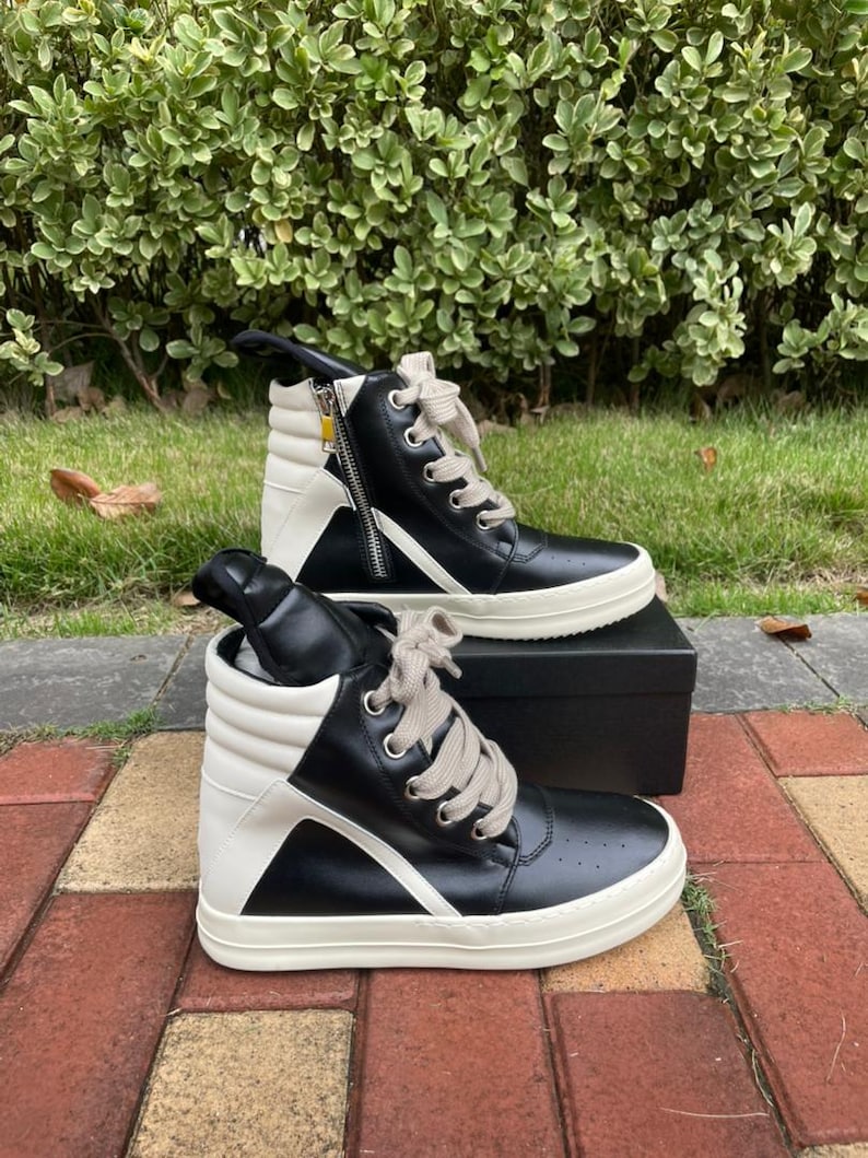 May include: Black and white high-top sneakers with a side zipper and light-colored laces. The sneakers have a thick, white sole and are set on a black box. The background features green foliage and a brick walkway.