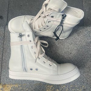 May include: Off-white high-top sneakers with side zippers and laces. The sneakers have a thick, white sole and a high ankle design. The shoes are made of a smooth material and feature silver-toned hardware.