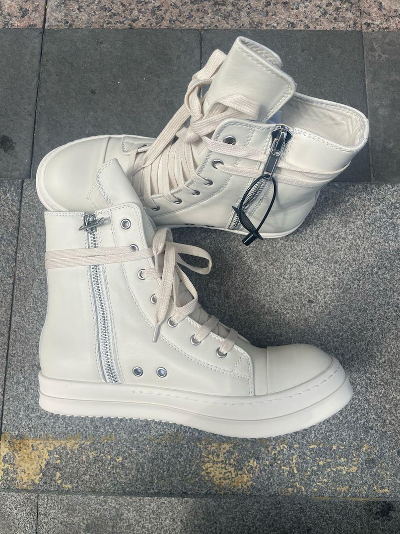 May include: Off-white high-top sneakers with side zippers and laces. The sneakers have a thick, white sole and a high ankle design. The shoes are made of a smooth material and feature silver-toned hardware.