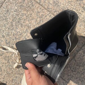 May include: A black high-top boot featuring a silver side zipper, white laces, and a black leather tongue with a grey and black graphic patch on the interior. The boot has a black interior lining and a white sole visible at the base of the shoe.