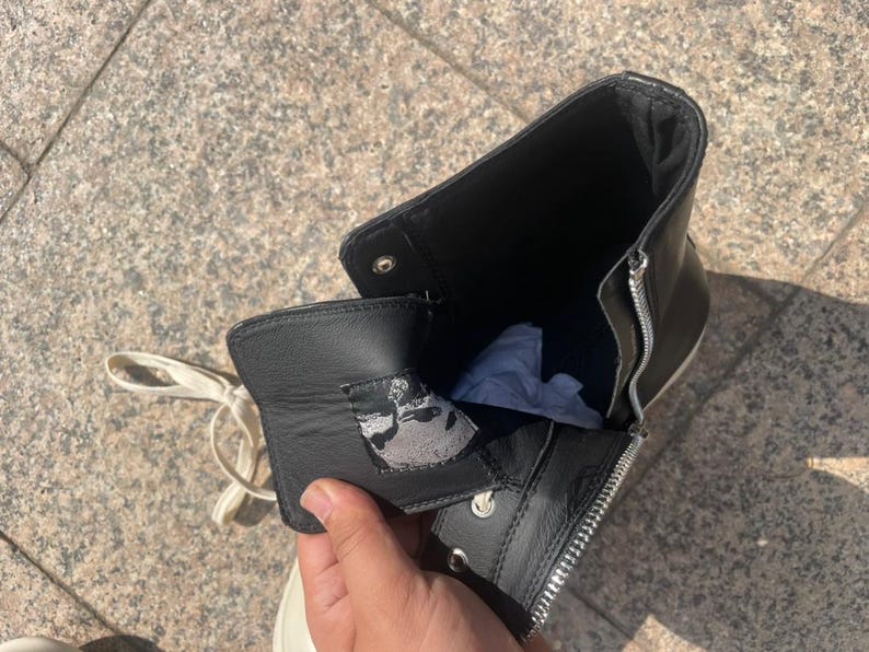 May include: A black high-top boot featuring a silver side zipper, white laces, and a black leather tongue with a grey and black graphic patch on the interior. The boot has a black interior lining and a white sole visible at the base of the shoe.