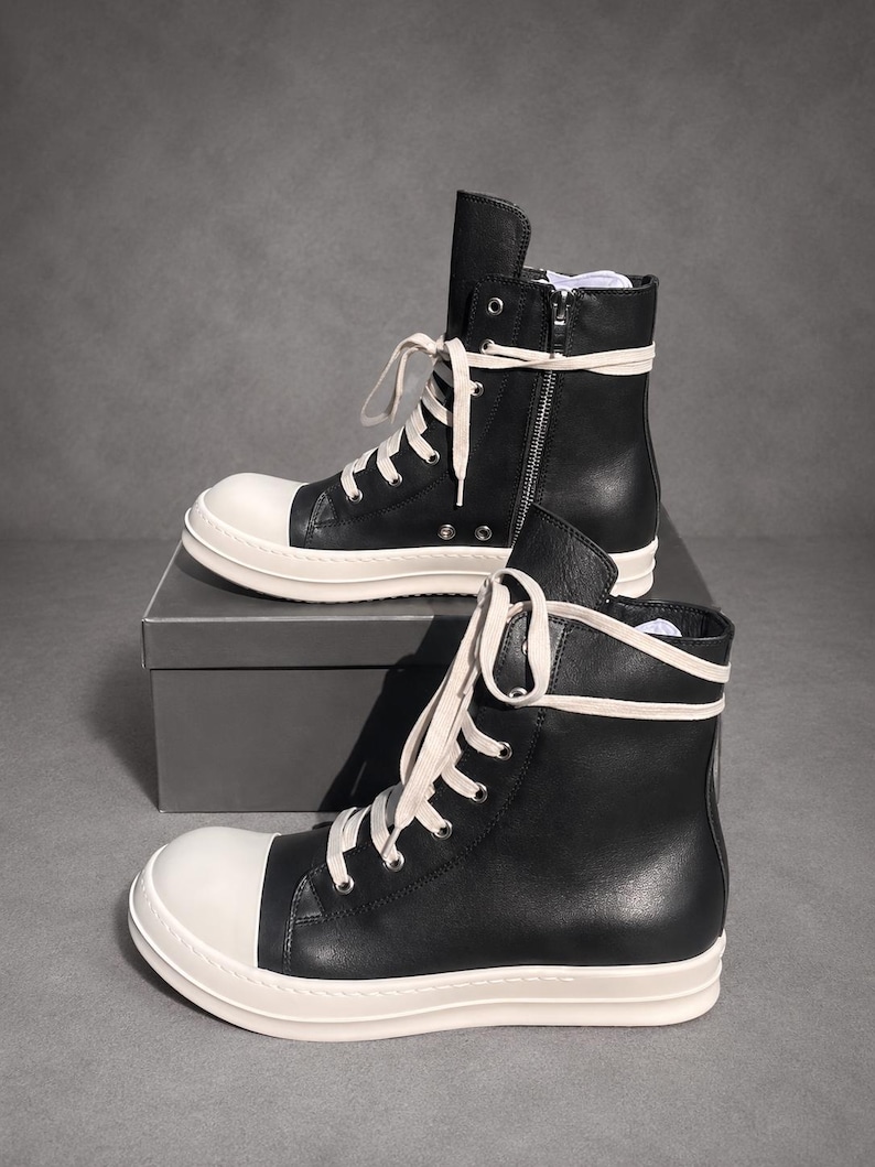 May include: A pair of high-top sneakers featuring black leather uppers, white rubber toe caps, and thick white rubber soles. The shoes include long, cream-colored laces threaded through silver-toned metal eyelets and a side zipper on the upper shoe.