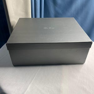 May include: A rectangular, gray box with a lid. The box has a matte finish and a subtle, embossed logo on the top. The box is sitting on a white surface, with a blue curtain in the background.