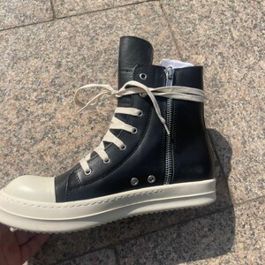 May include: A black high-top sneaker with a smooth leather upper, a prominent off-white rubber toe cap, and a matching thick rubber sole. The shoe features long cream-colored laces, silver-toned metal eyelets, and a vertical silver side zipper.