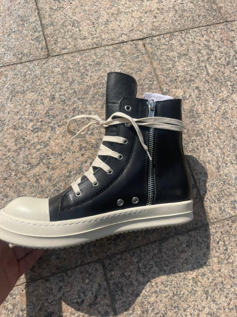 May include: A black high-top sneaker with a smooth leather upper, a prominent off-white rubber toe cap, and a matching thick rubber sole. The shoe features long cream-colored laces, silver-toned metal eyelets, and a vertical silver side zipper.