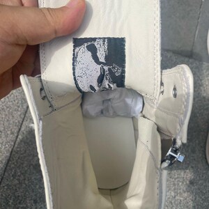 May include: Off-white high-top sneaker with a side zipper. The interior reveals a white insole and a black and white label with a skull design. The shoe is made of a smooth, leather-like material.