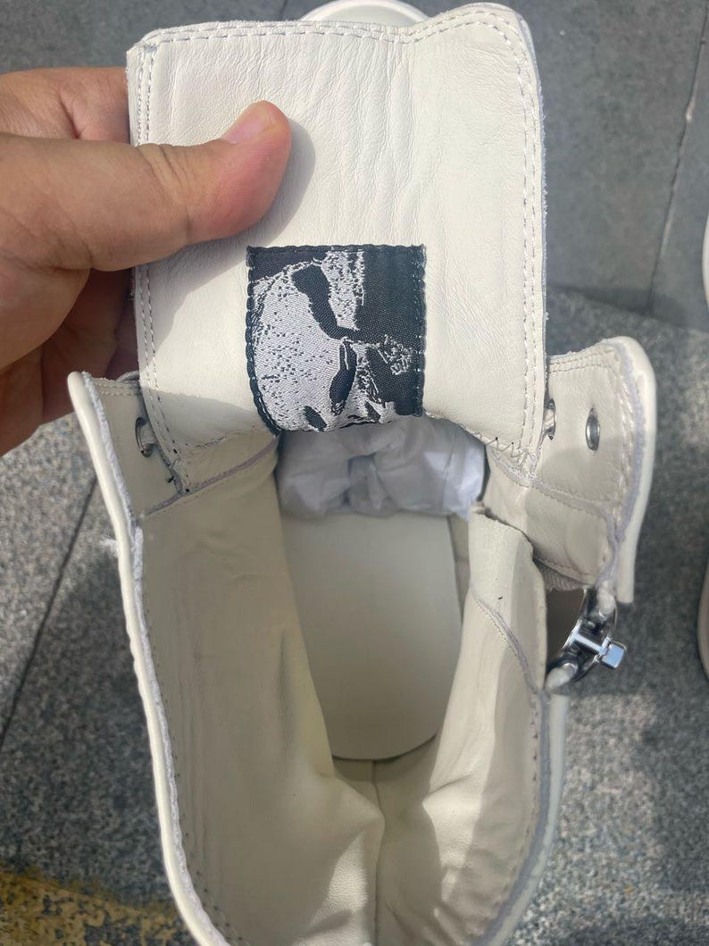 May include: Off-white high-top sneaker with a side zipper. The interior reveals a white insole and a black and white label with a skull design. The shoe is made of a smooth, leather-like material.