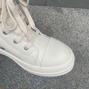 May include: Close-up of a white high-top sneaker with cream-colored laces. The shoe features a rounded toe, metal eyelets, and a thick, textured sole. The sneaker is made of a smooth, leather-like material.