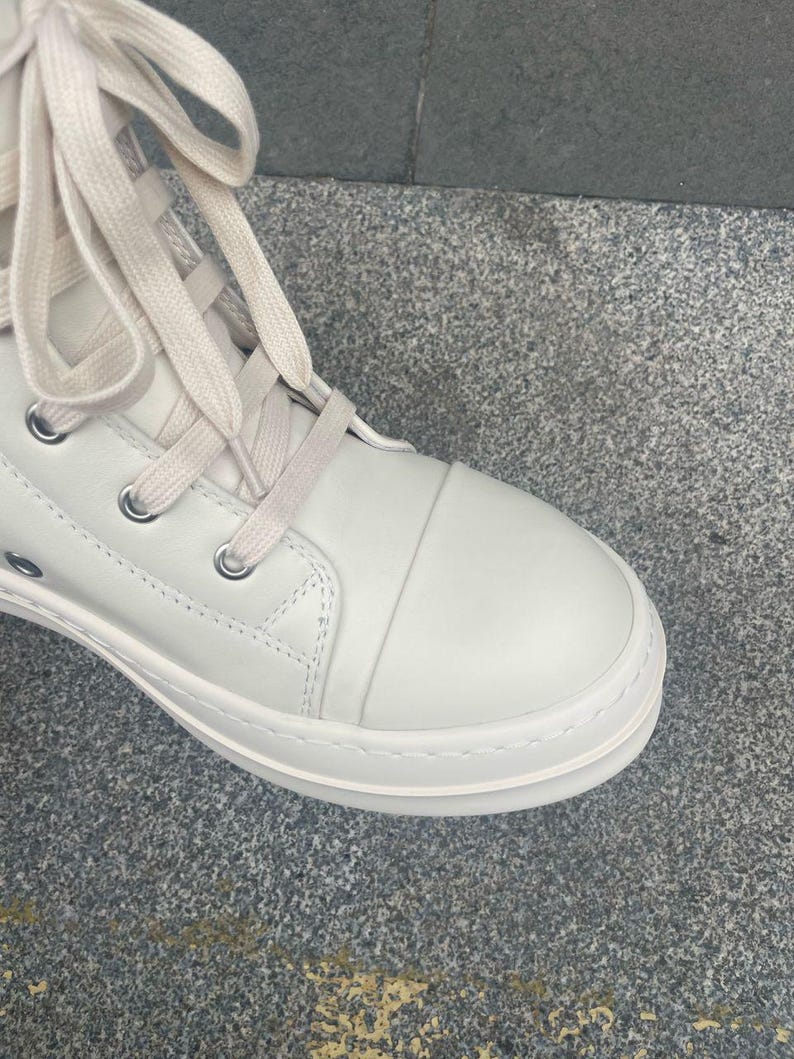 May include: Close-up of a white high-top sneaker with cream-colored laces. The shoe features a rounded toe, metal eyelets, and a thick, textured sole. The sneaker is made of a smooth, leather-like material.