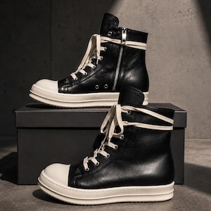 May include: Black high-top sneakers with white soles and laces. The sneakers have a zipper on the side and are displayed on a black box. The shoes have a minimalist design.