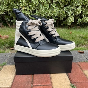 May include: A pair of high-top sneakers in black leather with white accents and a white sole. The sneakers have a unique design with a high tongue and are laced with beige laces. They are displayed on a black box.